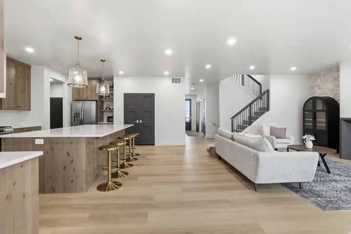 Open-concept living space featuring light wood-finish flooring, a kitchen island with a light countertop, recessed lighting, a staircase with dark metal railings, and a partial stone accent wall