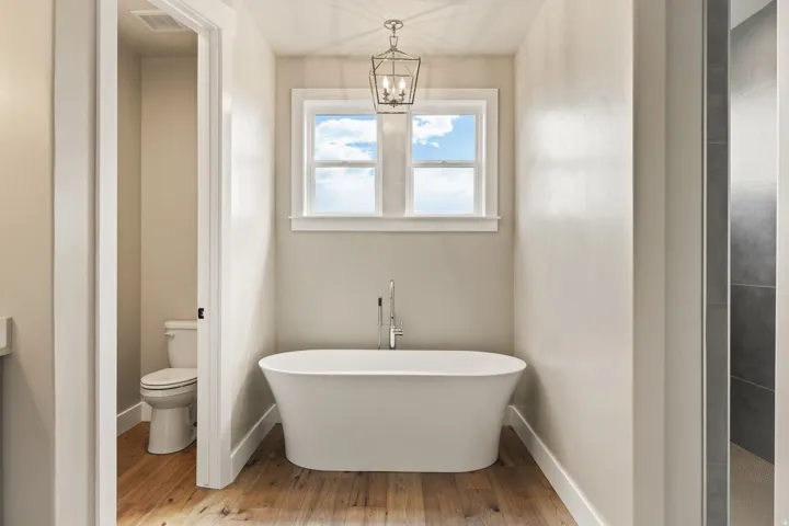Freestanding soaking tub with a floor-mounted faucet, wood-finish flooring, and a double-pane window