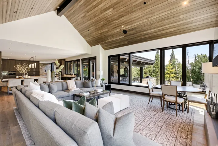 Spacious open-concept living area featuring a vaulted wood-finish ceiling, large black-framed picture windows showcasing exterior greenery, and wood-finish flooring