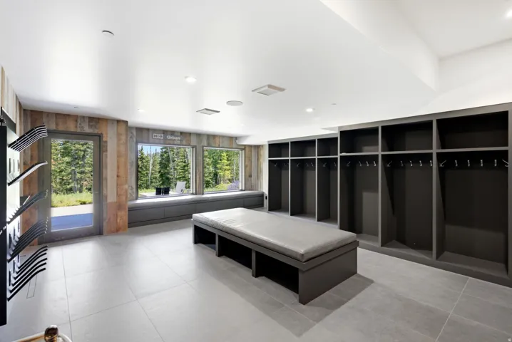 Spacious room featuring light gray tile flooring, extensive built-in open lockers with hanging rods, a central upholstered bench, and a wall of windows showcasing natural scenery