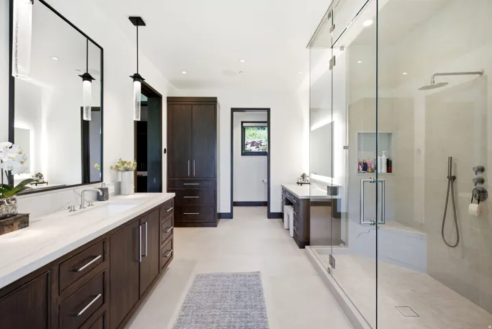 Spacious bathroom featuring a frameless glass shower enclosure with a built-in bench, dual vanity with a white countertop, dark wood-finish cabinetry, and recessed lighting