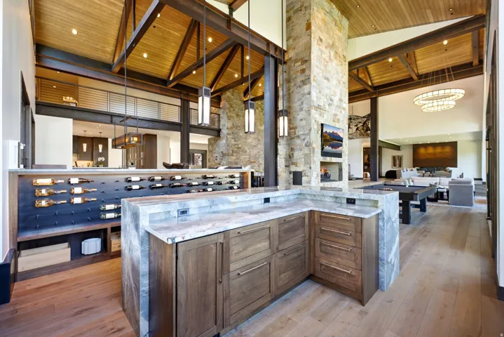 Vaulted wood-paneled ceilings with exposed beams, stone-clad fireplace with built-in media, integrated wine storage, light wood-finish flooring, and a built-in bar with stone countertops and wood cabinetry