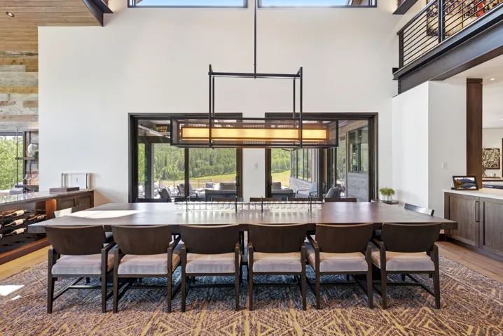 Spacious dining area featuring a vaulted ceiling with exposed beams, an expansive window wall, a linear chandelier, and a built-in wine rack