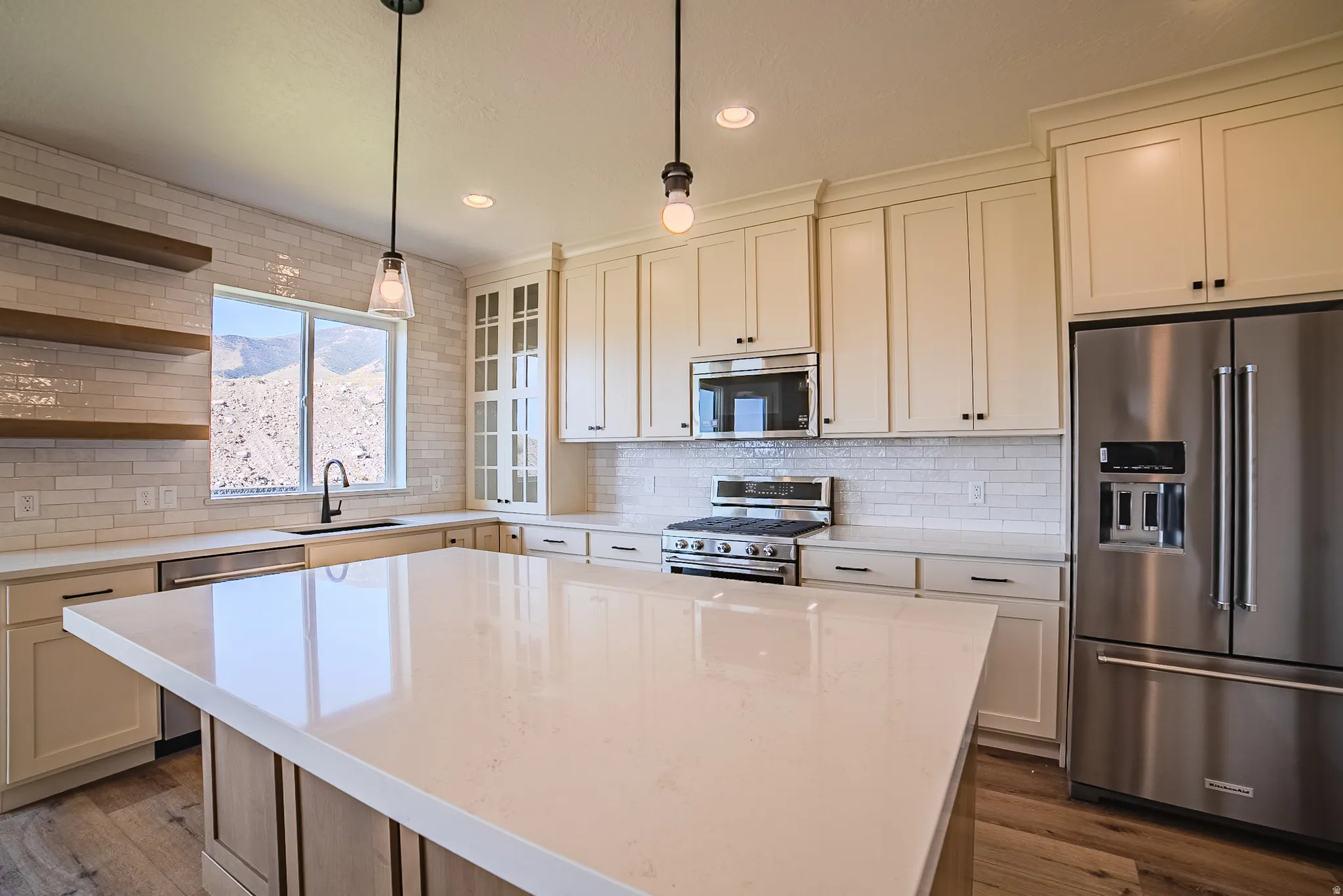 Spacious kitchen featuring a large central island with a light-toned countertop, extensive white subway tile backsplash, stainless steel appliances, ivory shaker-style cabinetry, and wood-finish flooring