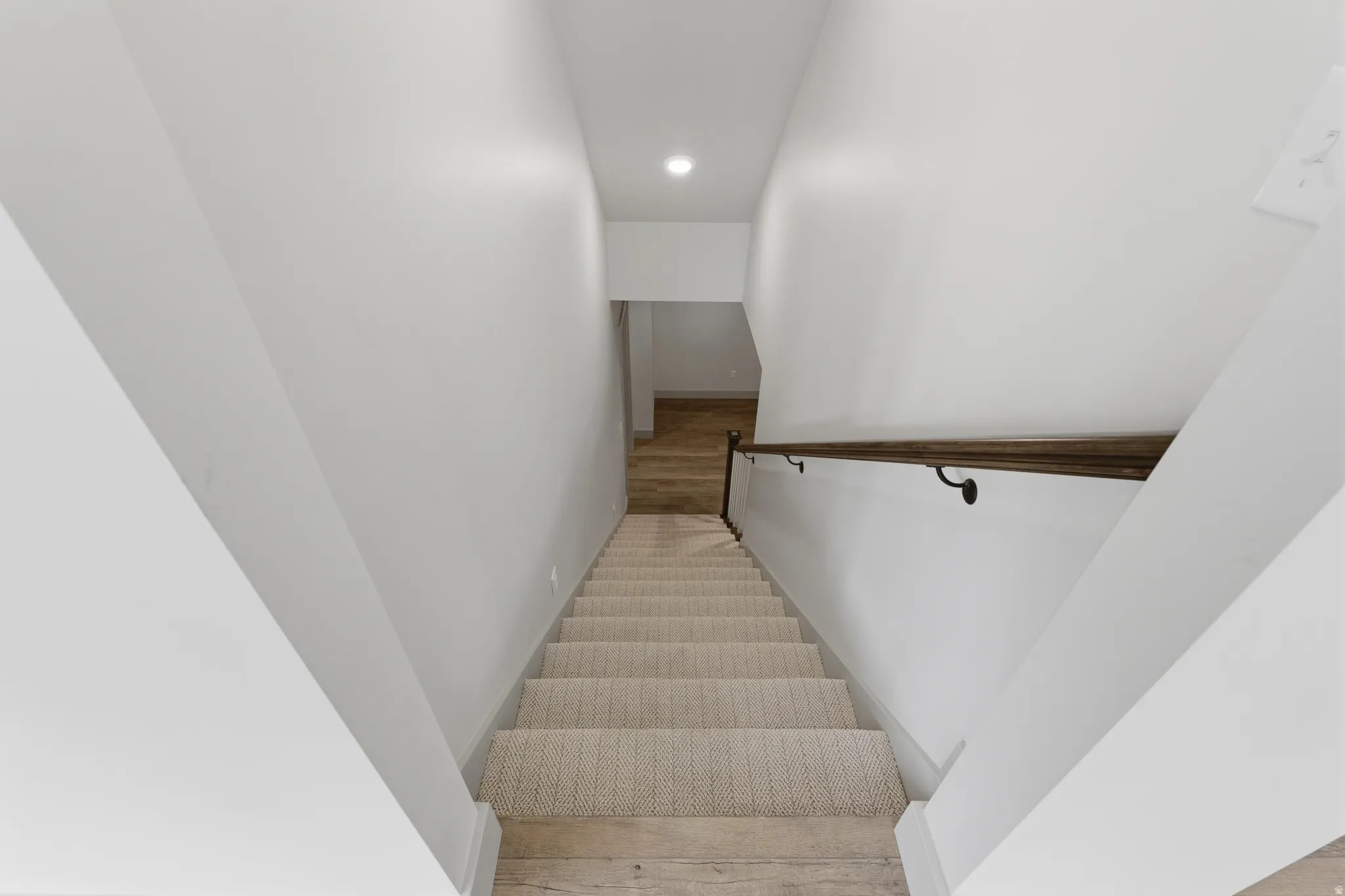 Staircase with light-toned carpeted treads, wood-finish flooring at the base, and a dark wood handrail with matte black brackets