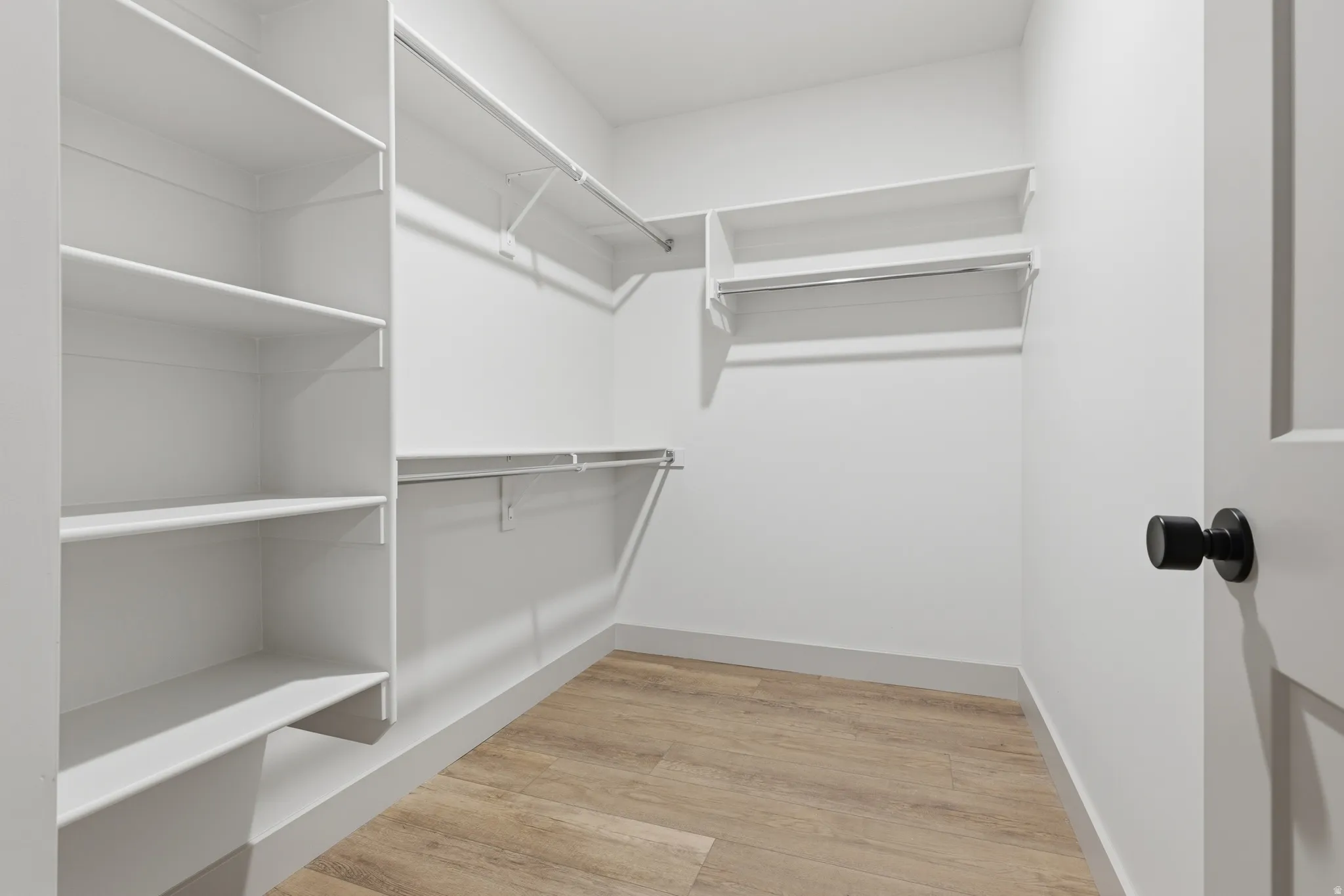 Walk-in closet featuring built-in shelving, multiple hanging rods, light wood-finish flooring, and a modern black door handle