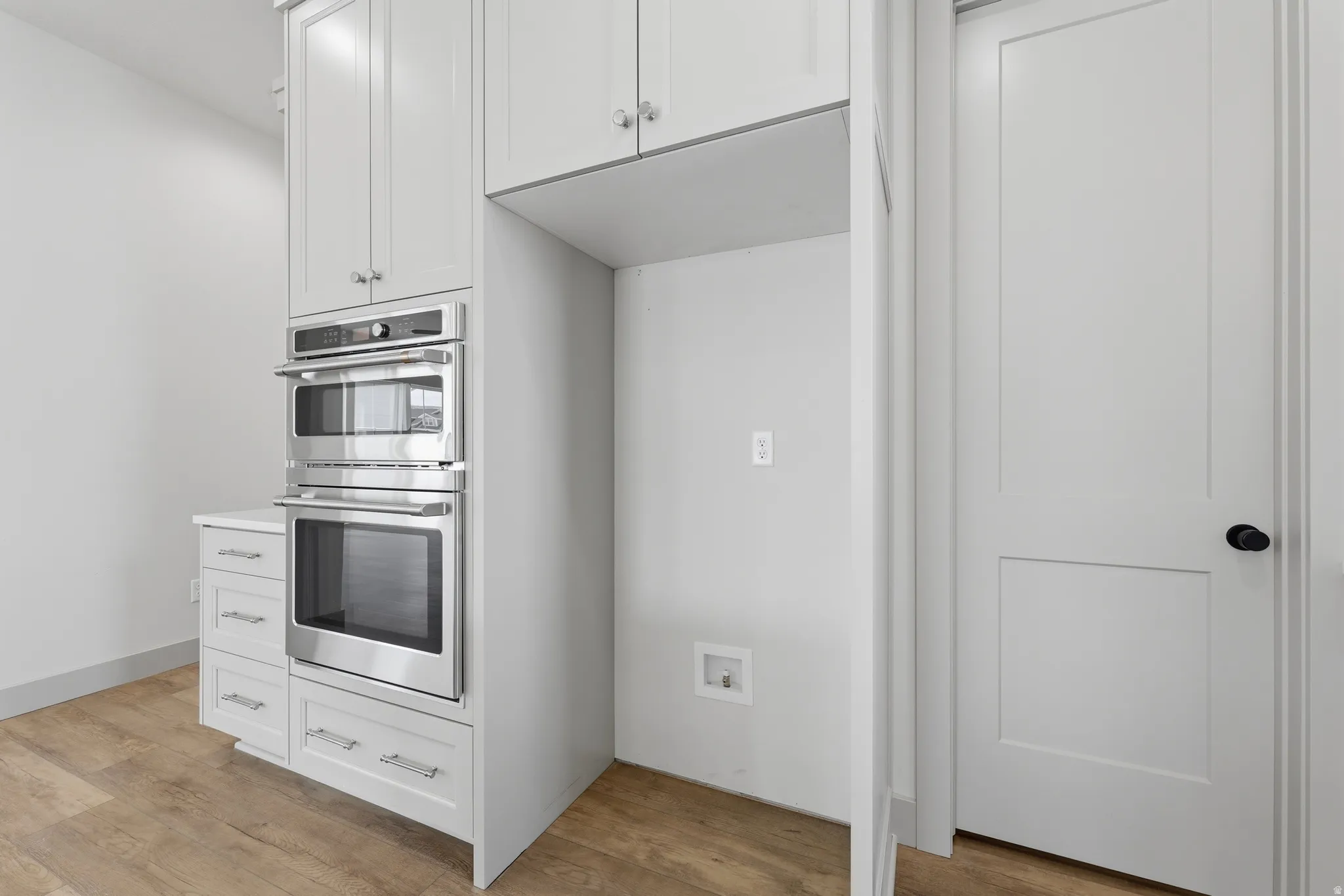 White shaker cabinetry with chrome hardware, integrated stainless steel double oven, wood-finish flooring, and a white recessed panel door