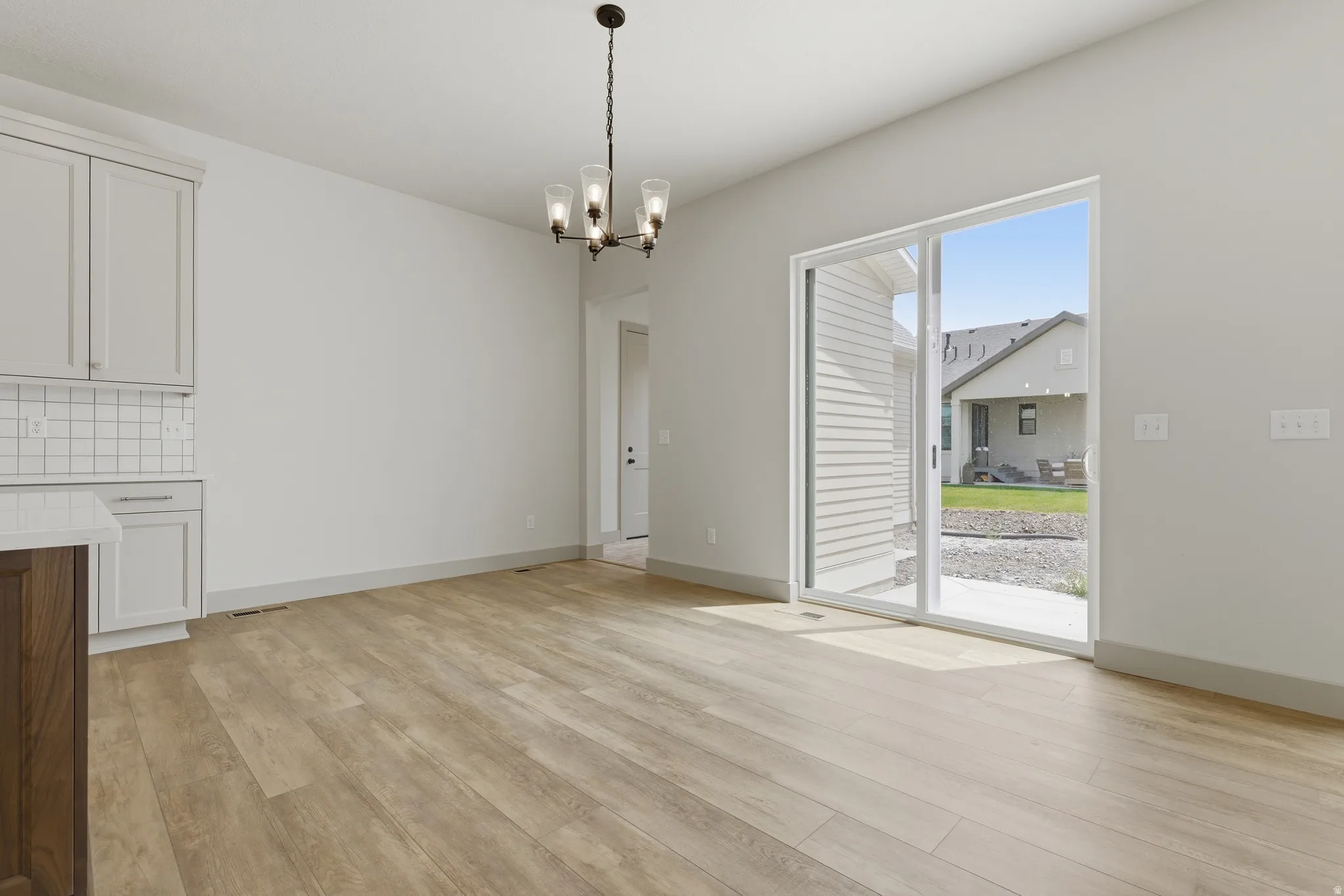 Spacious room with wood-finish flooring, featuring a modern chandelier, light-toned cabinetry with a subway tile backsplash, and a large sliding glass door