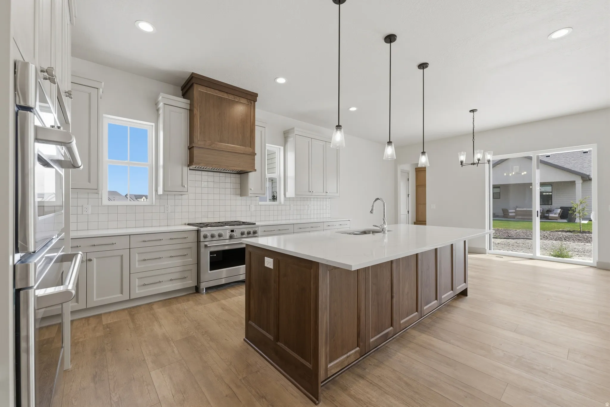 Modern kitchen featuring a large central island with a sink and pendant lighting, light wood-finish flooring, light gray cabinetry, stainless steel appliances, and subway tile backsplash
