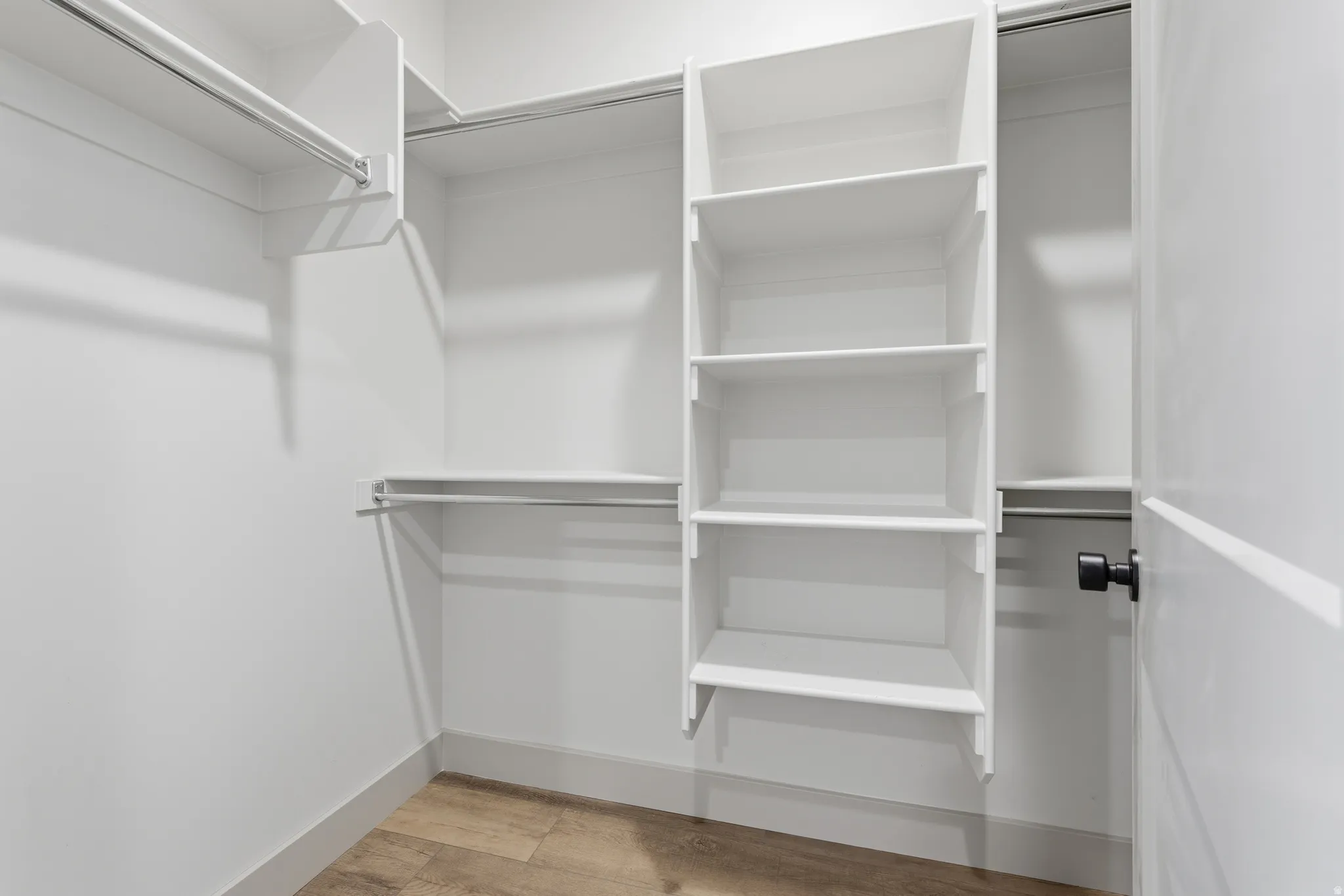 Spacious walk-in closet featuring built-in shelving, multiple hanging rods, white walls, light wood-finish flooring, and a white door with matte black hardware
