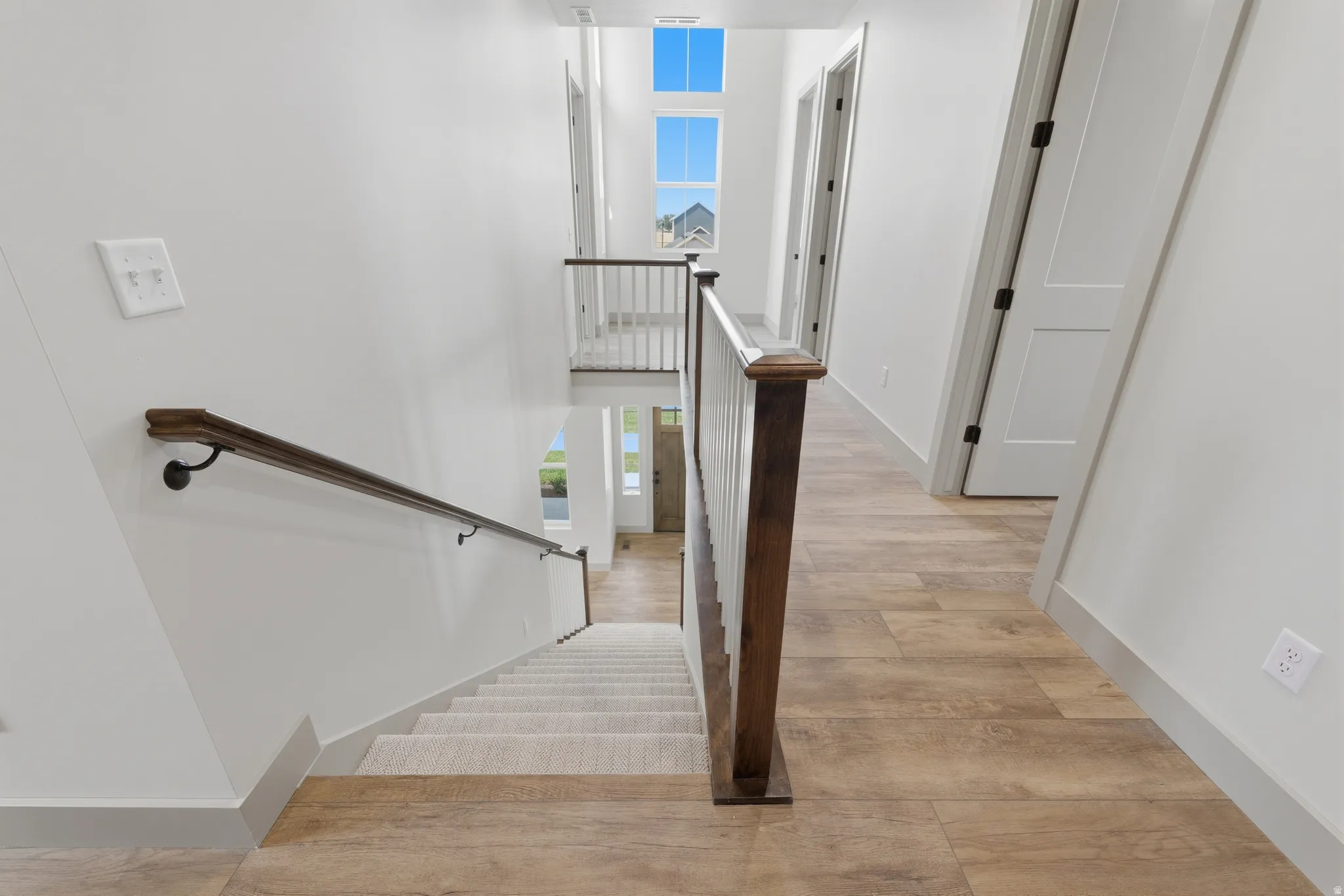 Staircase with carpeted treads and a dark wood handrail, featuring a white balustrade overlooking a bright entry foyer
