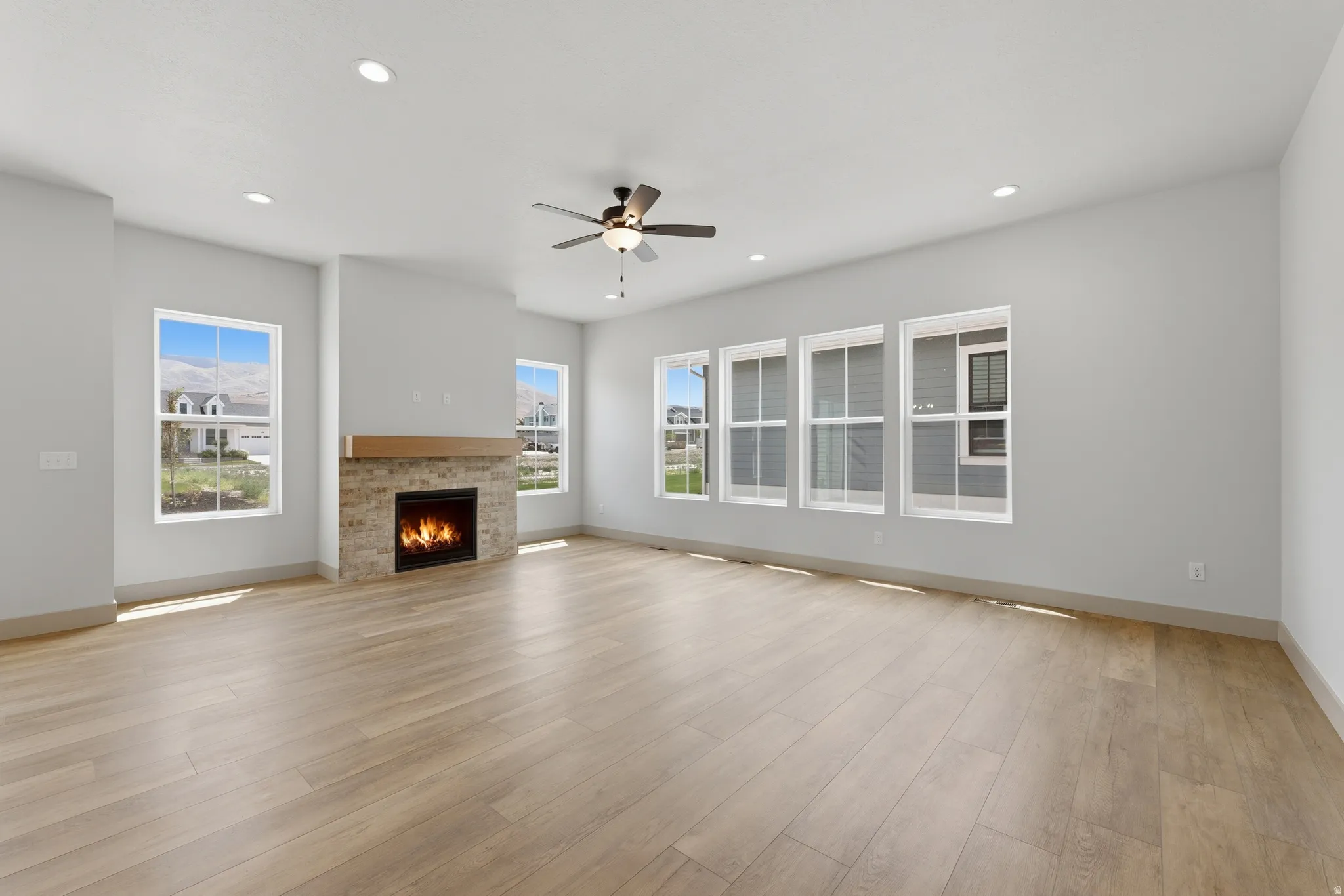 Spacious living area featuring wood-finish flooring, a stone-clad fireplace with a wood mantel, multiple windows, recessed lighting, and a ceiling fan