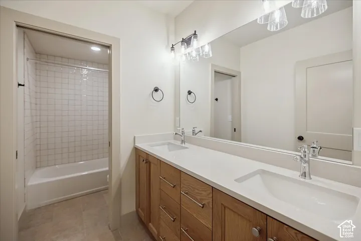 Bathroom featuring a dual-sink wood-finish vanity with a stone countertop, wall-to-wall mirror, polished metal fixtures, and a tile-surround shower and tub combination