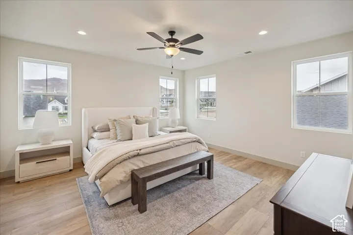 Primary Suite. Neutral-toned bedroom featuring wood-finish flooring, recessed lighting, a central ceiling fan with integrated lighting, and multiple windows with muntin detailing