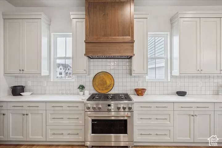 Kitchen featuring custom white cabinetry, stone countertops, square tile backsplash, stainless steel gas range, and a wood-finish hood vent