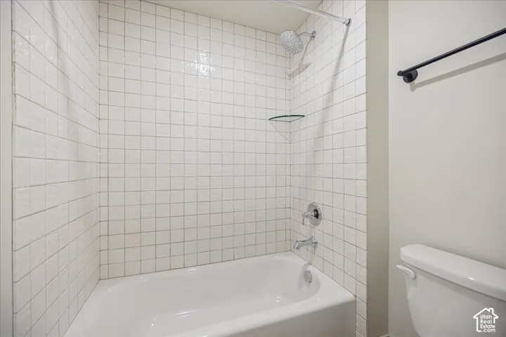 Bathtub and shower combination featuring a white tiled surround, chrome shower fixtures, integrated glass shelving, and a wall-mounted towel bar