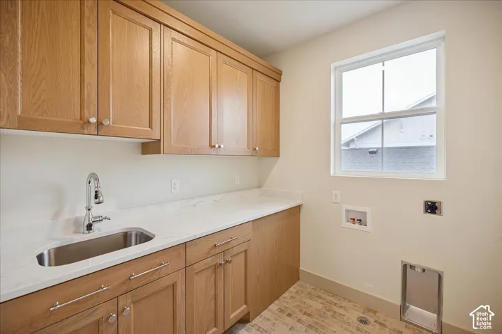 Laundry space featuring wood-finish cabinetry, light-toned stone-style countertop, undermount stainless steel sink with pull-down faucet, and neutral tile flooring