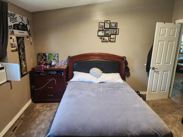 Carpeted room featuring a wood-finish headboard with an integrated upholstered panel, a multi-paneled interior door, and a wall-mounted vent