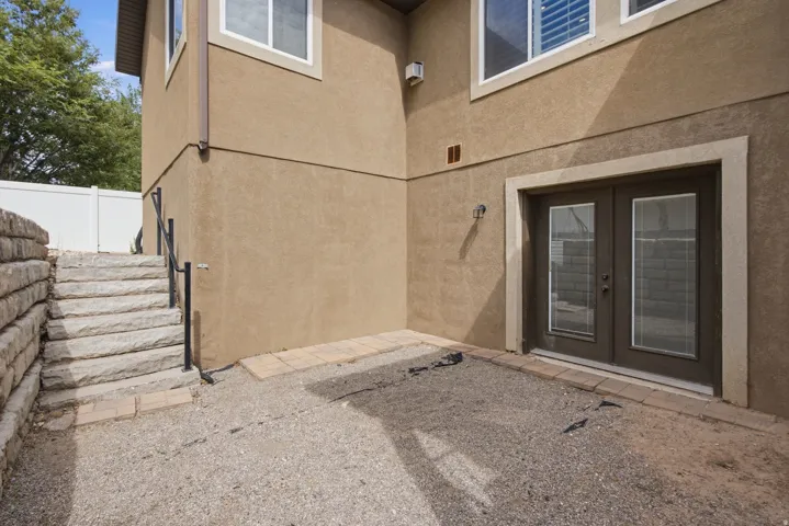 Ground-level patio area featuring double French doors with integrated blinds, a stucco exterior, and stone-retaining wall with matching stairs and black metal handrails