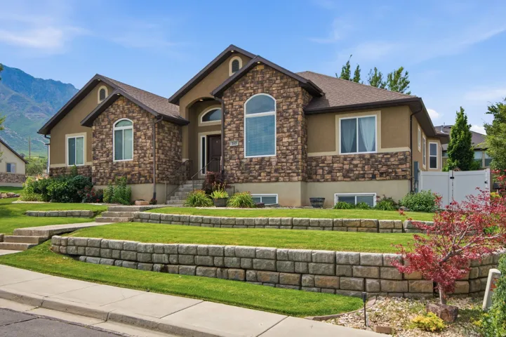 Stately residence featuring a stone and stucco exterior, arched windows, tiered landscaping with retaining walls, a mature shrub border, and a prominent front entry with steps
