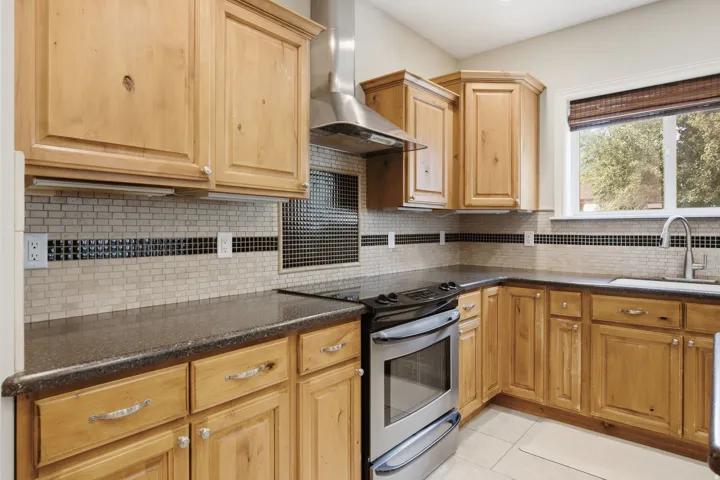 Kitchen featuring wood-finish cabinetry, dark countertops, a stainless steel range with matching range hood, and a tile backsplash with decorative accents