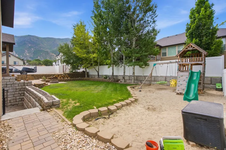 Expansive backyard featuring a manicured lawn, a sand play area, paver stone patio, tiered retaining walls, and mature trees