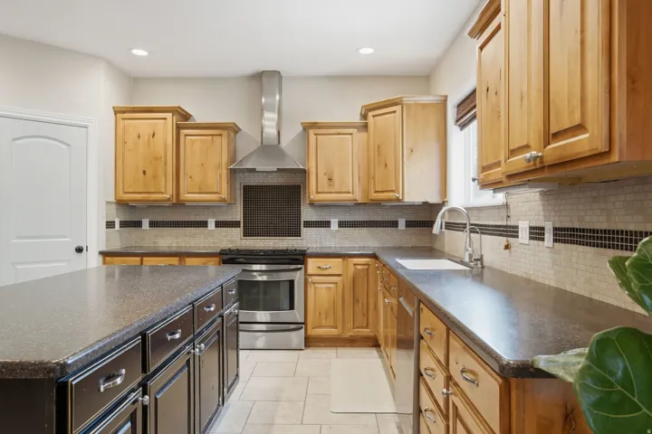 Kitchen featuring an island with dark cabinetry, light wood-finish upper and lower cabinetry, stone-look countertops, stainless steel appliances, and a mosaic tile backsplash