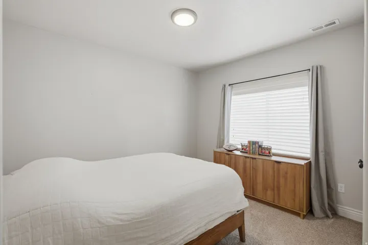 Bedroom with light gray walls, carpet flooring, and a large window with white blinds and drapes