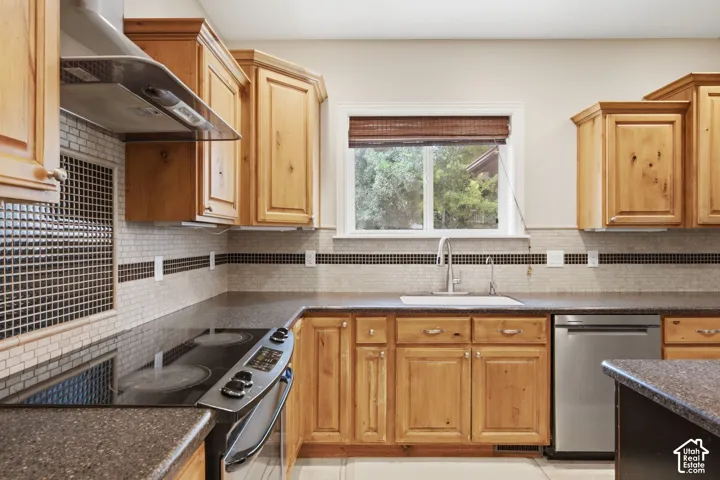 Kitchen featuring wood cabinetry, dark countertops, tile backsplash with accent strip, stainless steel appliances, and a window above the sink