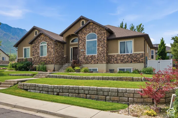 Stately residence featuring a stone and stucco exterior, arched windows, tiered landscaping with retaining walls, a mature shrub border, and a prominent front entry with steps