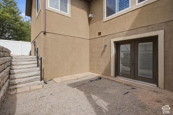 Ground-level patio area featuring double French doors with integrated blinds, a stucco exterior, and stone-retaining wall with matching stairs and black metal handrails