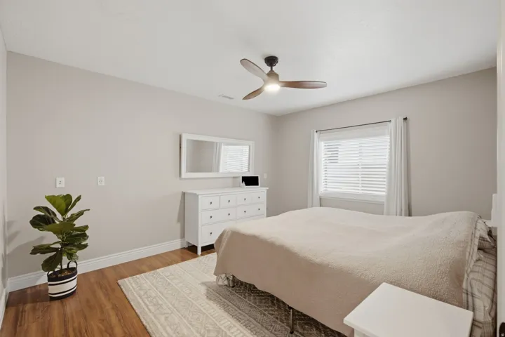 Spacious room featuring wood-finish flooring, a ceiling fan with integrated lighting, and a window with white blinds and curtains