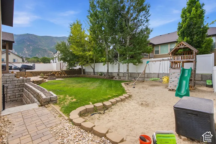 Expansive backyard featuring a manicured lawn, a sand play area, paver stone patio, tiered retaining walls, and mature trees