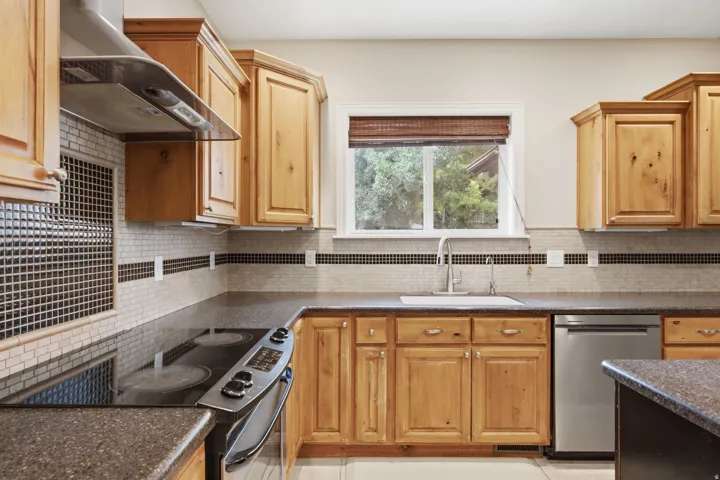 Kitchen featuring wood cabinetry, dark countertops, tile backsplash with accent strip, stainless steel appliances, and a window above the sink
