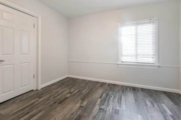 Spacious room featuring wood-finish flooring, light neutral wall tones, a window with blinds, white trim, and a paneled door