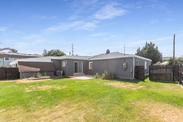 Spacious backyard featuring a large green lawn, a concrete patio, dark gray painted brick siding, a sliding glass door, and dark wood fencing