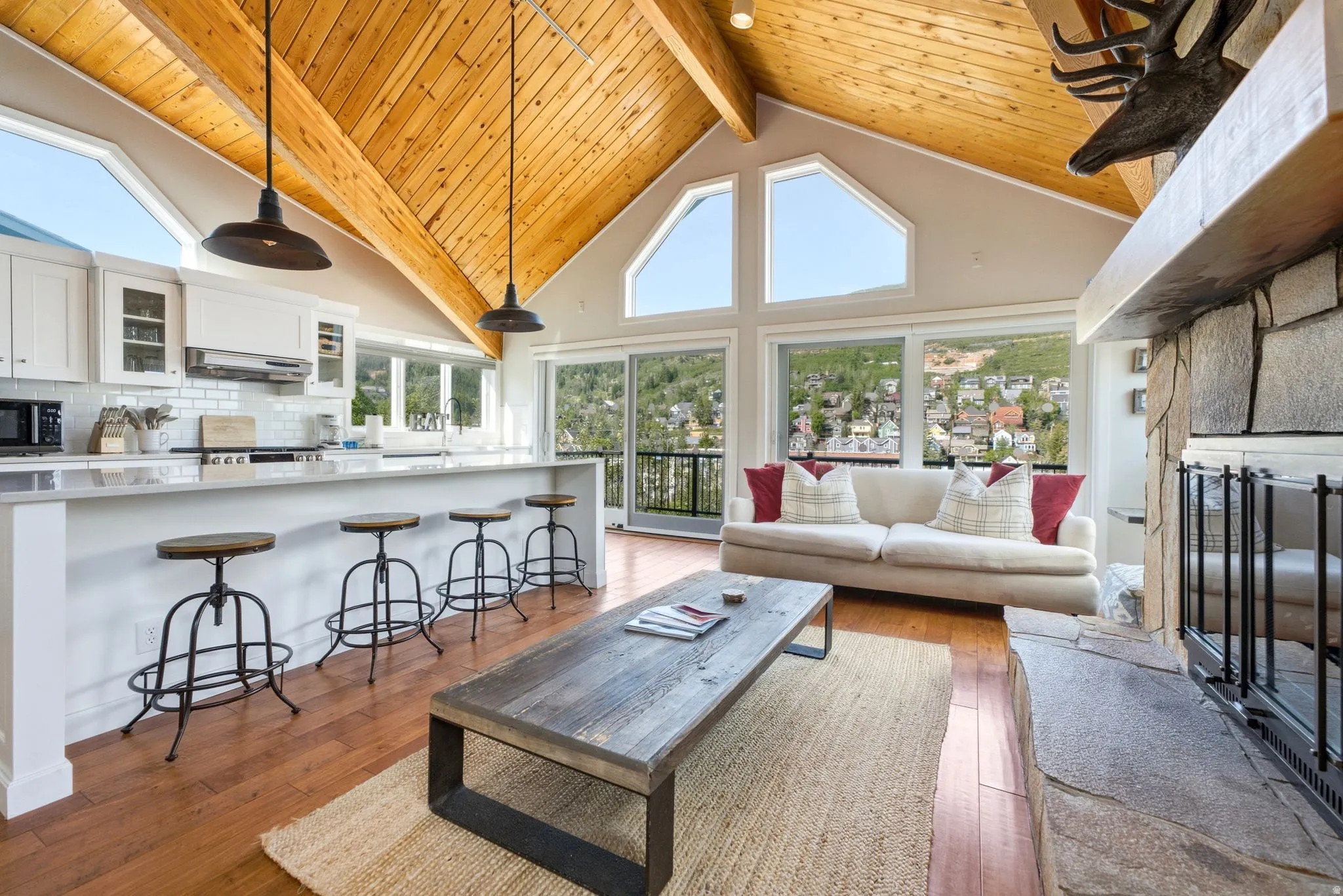 Open-concept living area featuring a vaulted wood-paneled ceiling with exposed beams, wood-finish flooring, a stone fireplace, and an integrated kitchen with white cabinetry and a breakfast bar