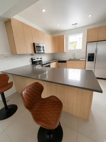 Modern kitchen featuring an expansive island with a solid surface countertop, light wood-finish cabinetry, stainless steel appliances, a tile backsplash, and recessed lighting