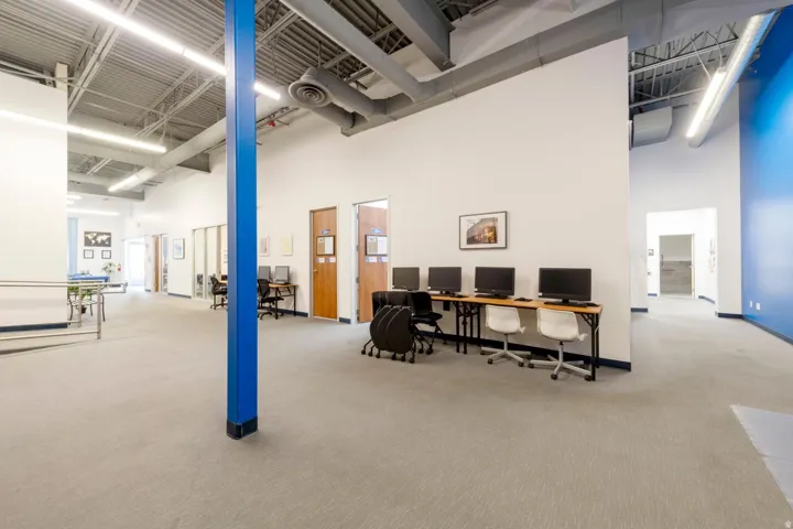 Spacious interior featuring a high exposed ceiling with linear fluorescent lighting, light gray carpeted flooring, and a dedicated computer workstation area