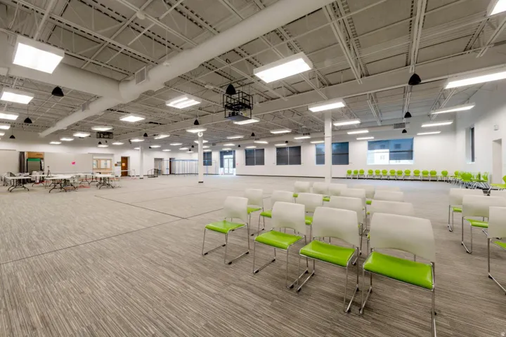 Expansive commercial space featuring high ceilings with exposed ductwork, fluorescent lighting, multiple windows, and a large carpeted floor area