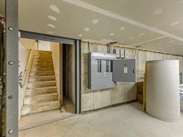 Basement space with concrete flooring and walls, featuring an exposed electrical panel, a white cylindrical tank, and a wooden staircase with a handrail