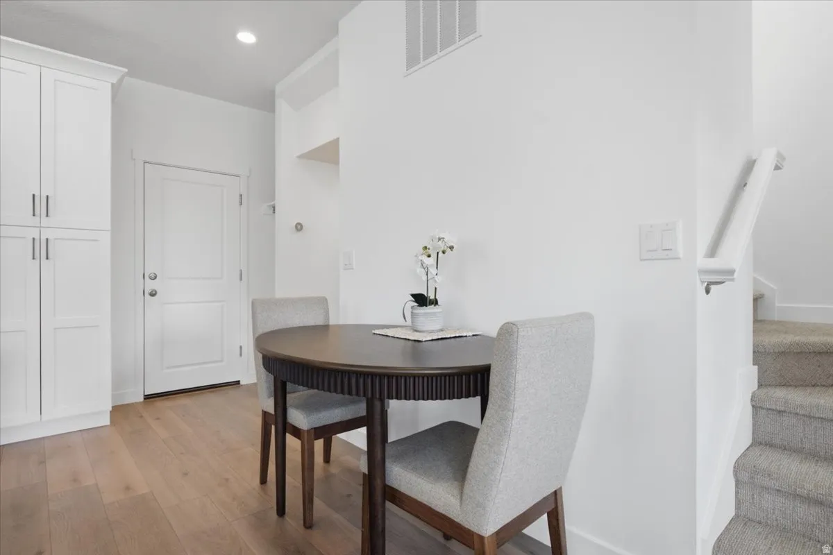 Bright, open foyer featuring warm wood-finish flooring, white shaker-style cabinetry with chrome hardware, a dark wood dining table with upholstered chairs, recessed ceiling lighting, and direct access to carpeted stairs