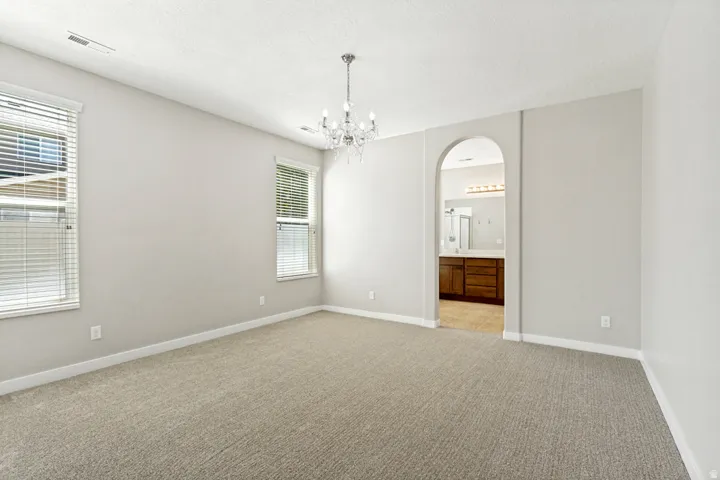 Carpeted primary bedroom featuring a crystal chandelier, two windows with blinds, and an arched doorway opening to a bathroom with a wood-finish vanity and mirror