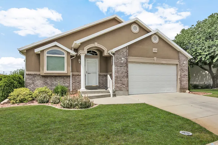 Exterior facade featuring a two-car garage, brick accents, stucco siding, a covered entry with white columns, and a manicured lawn with established landscaping