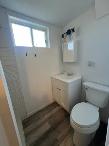 White vanity with an integrated sink, wood-finish flooring, white toilet, and a window providing natural light