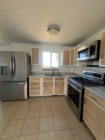 Kitchen featuring stainless steel appliances, tile flooring, wood-finish cabinetry, light-toned countertops, and a window above the sink
