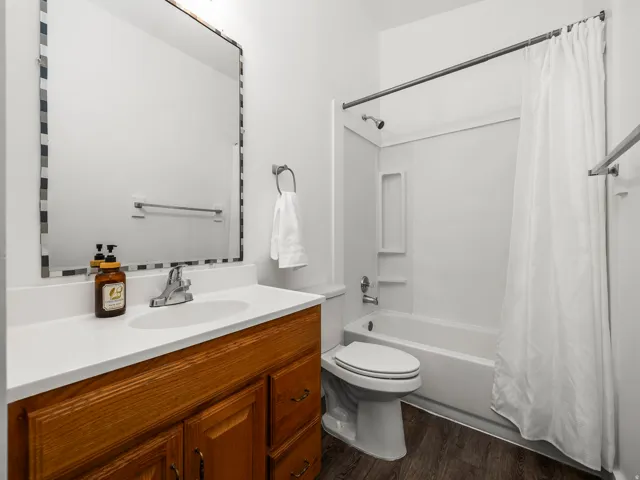 Bathroom featuring a wood-finish vanity with an integrated sink and white countertop, a mirror with a decorative tile frame, a shower/tub combination with white surround, and dark wood-finish flooring