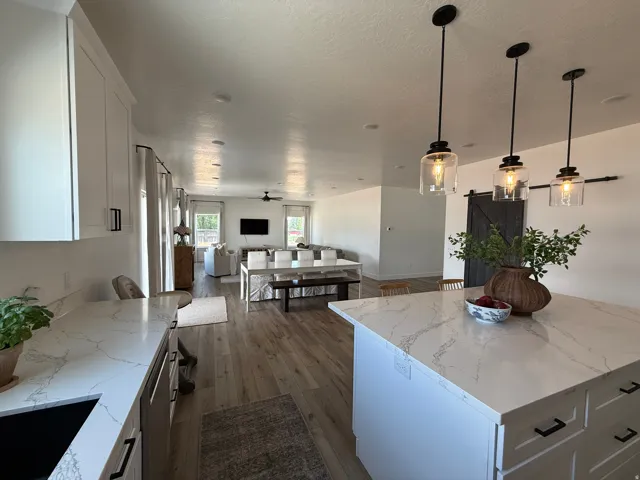 Open-concept living space featuring a spacious kitchen island with a light-toned countertop, three pendant lights, wood-finish flooring, white cabinetry with black hardware, and a dark wood barn door