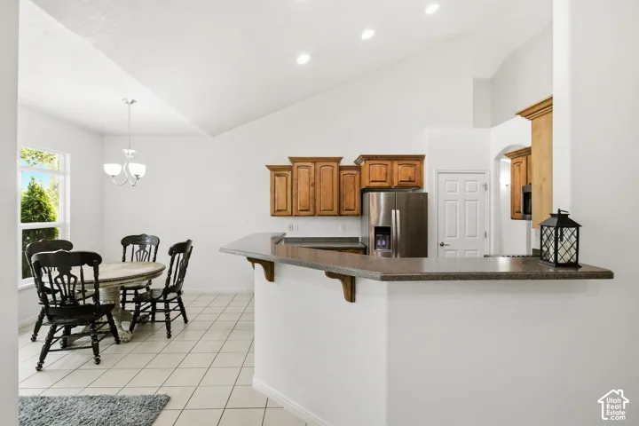 Open-concept kitchen and dining area featuring a vaulted ceiling, recessed lighting, wood cabinetry, stainless steel refrigerator, and a breakfast bar with a stone-finish countertop