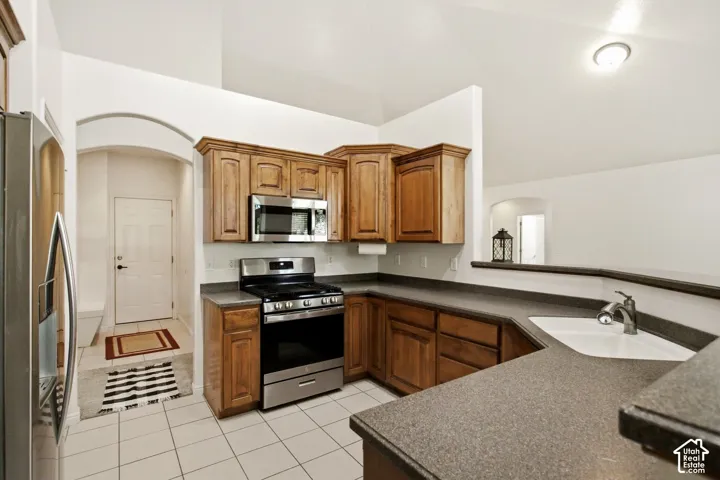 Kitchen featuring wood cabinetry, stainless steel appliances, a corner sink with a gooseneck faucet, composite countertops, and white tile flooring